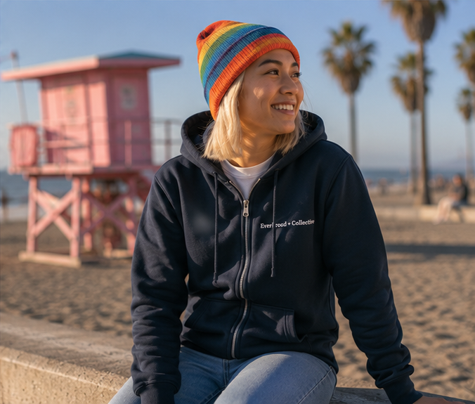EverProud Logo Minimalist Zip Hoodie on a bright Pacific Northwest coastal beach — queer-owned PNW Pride apparel, front view