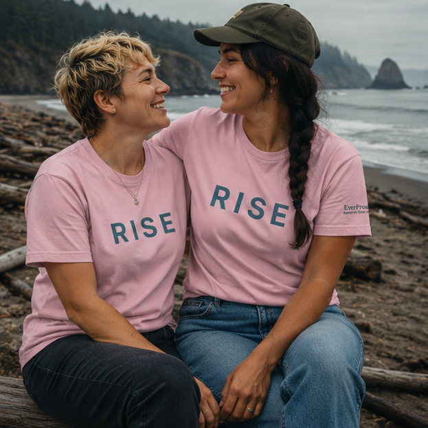 Rise Women's Relaxed T-Shirt on a misty Pacific Northwest beach — queer-owned PNW Pride apparel, front view